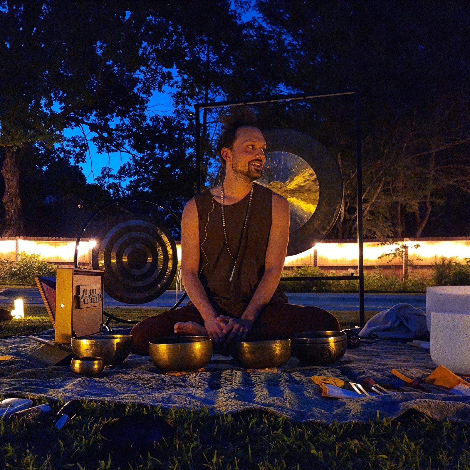 Matias sitting on a blanket outdoors at dusk among singing bowls, two gongs lit behind him