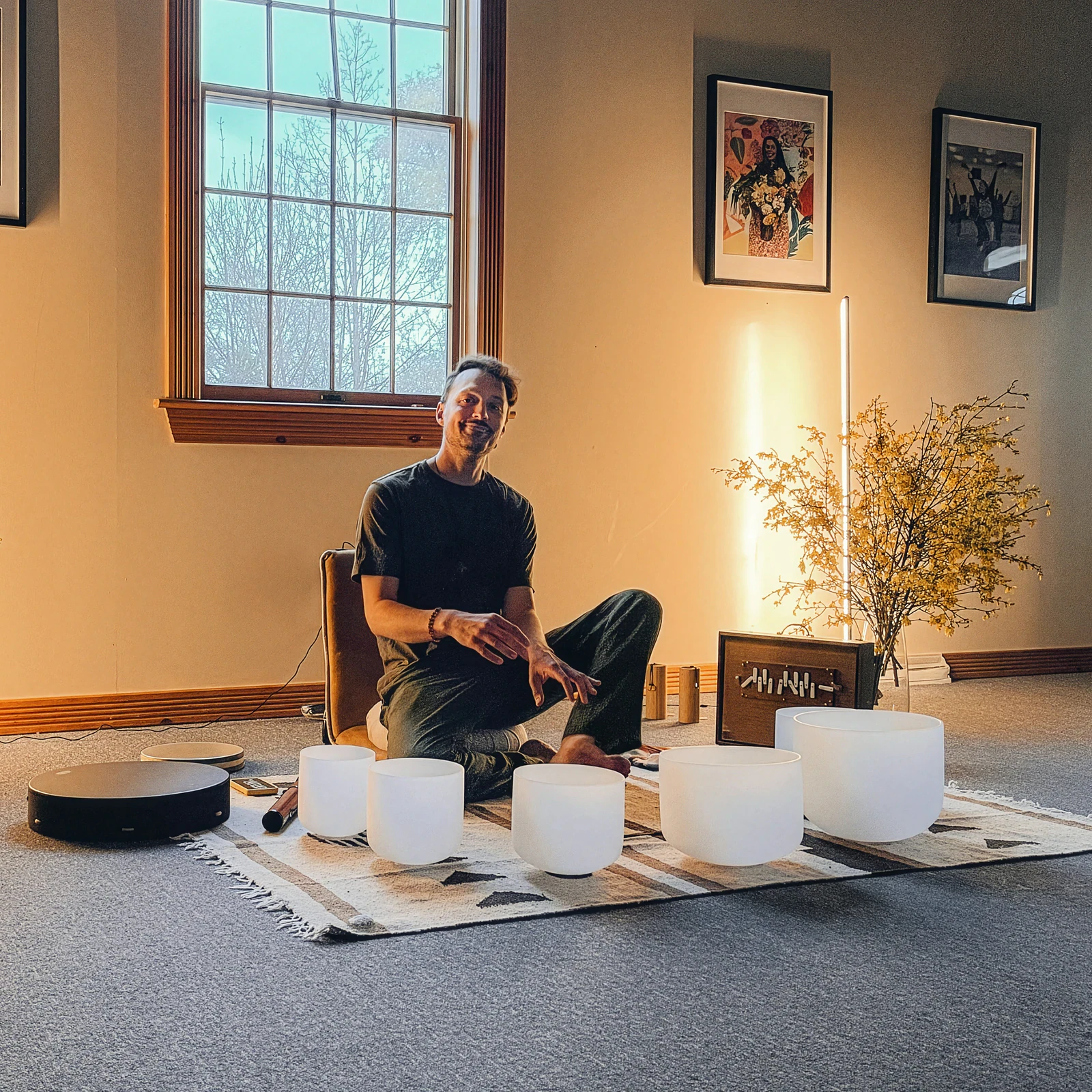 Matias smiling, sitting cross-legged on a rug in a warmly lit room behind a row of crystal singing bowls