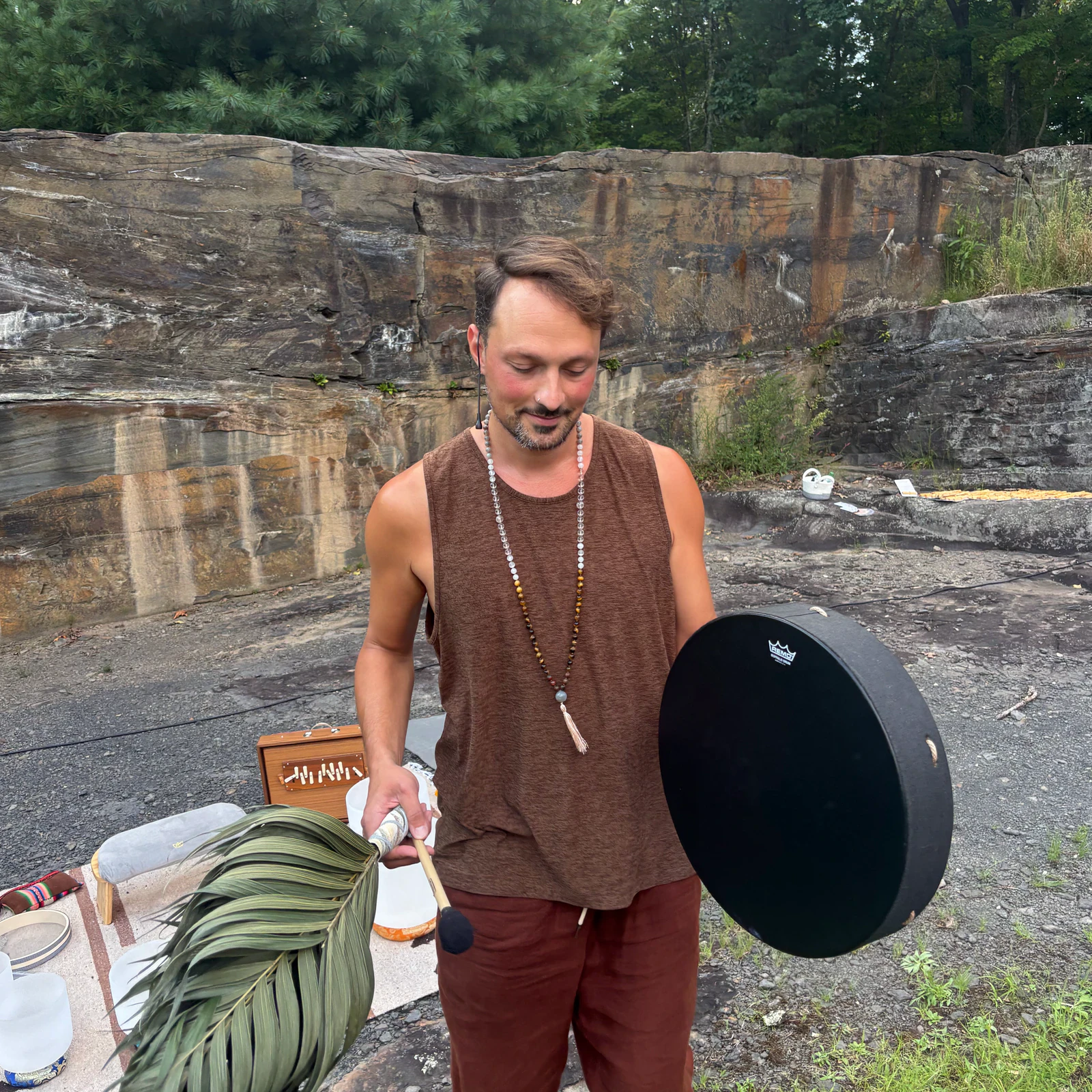 Matias outdoors in front of a rock face, holding a frame drum and mallet, a palm frond and a sound bath set up beside him